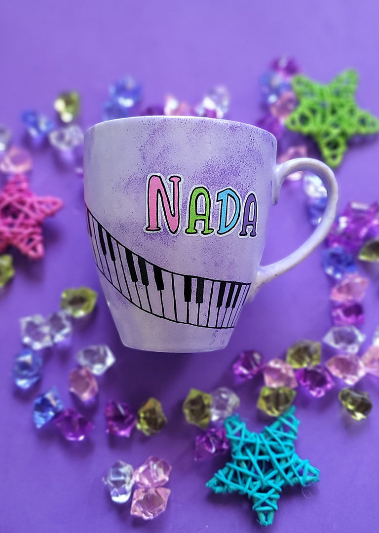 A Lilac hand painted mug with the name nada and piano buttons drawn diagonally on the mug