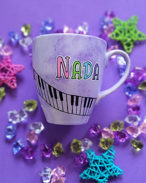 A Lilac hand painted mug with the name nada and piano buttons drawn diagonally on the mug