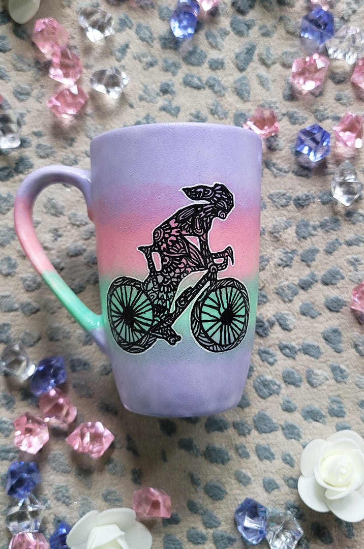 A gradient hand painted mug (lilac-pink-mint-lilac) with a silhouette of a girl on a bike drawn and filled with patterns