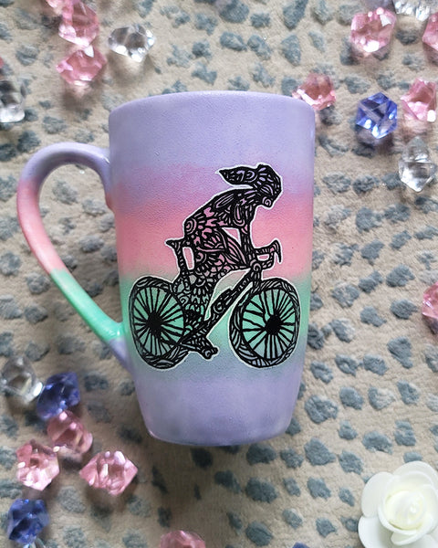 A gradient hand painted mug (lilac-pink-mint-lilac) with a silhouette of a girl on a bike drawn and filled with patterns