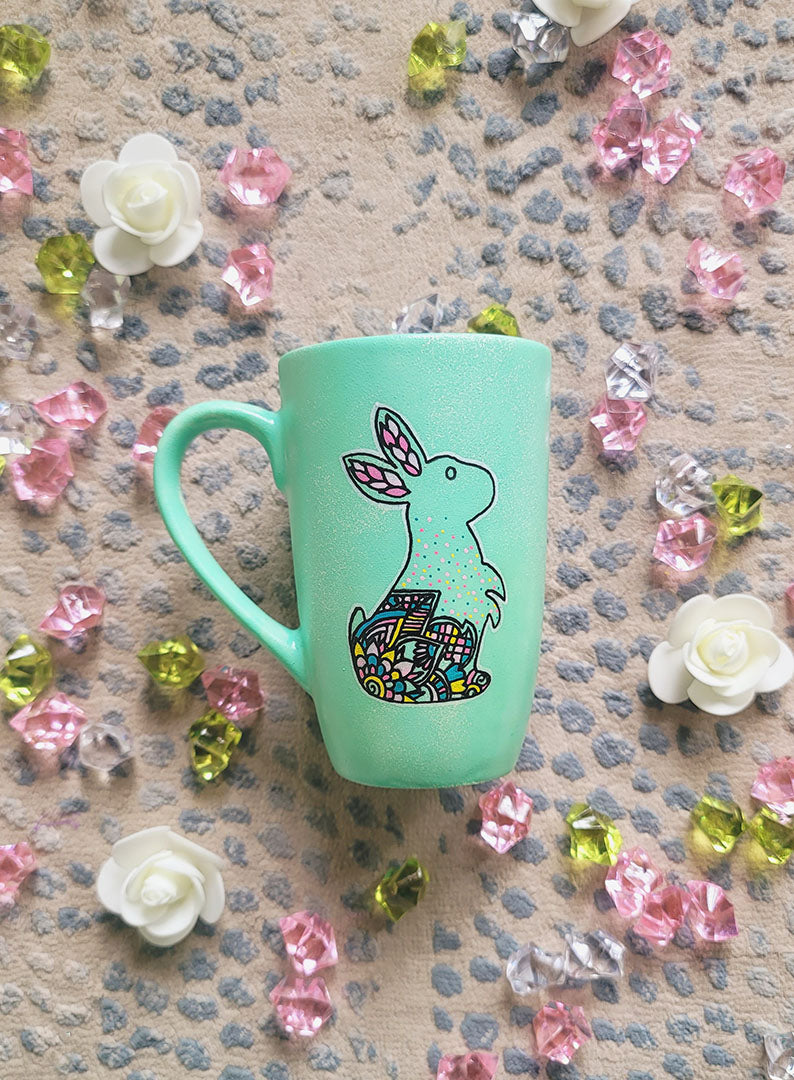 A Mint Hand Painted mug with a bunny drawn on it in black looking up and the bunny itself  has patterns on the corner inside it