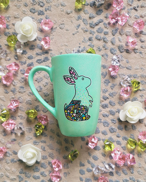 A Mint Hand Painted mug with a bunny drawn on it in black looking up and the bunny itself  has patterns on the corner inside it