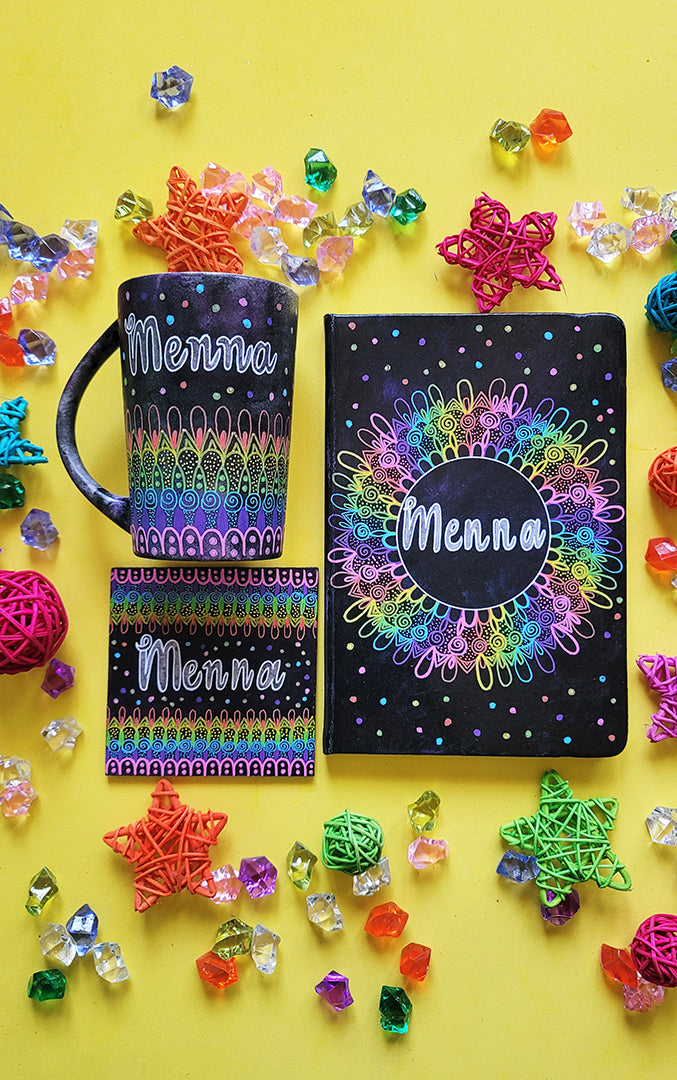 A Dark Sky hand painted mug, notebook and coaster with patterns on the bottom half horizontally inlayers of pink then purple then blue then green then yellow then orange, with colored dots scattered all over and the name "Menna" written in silver on the top