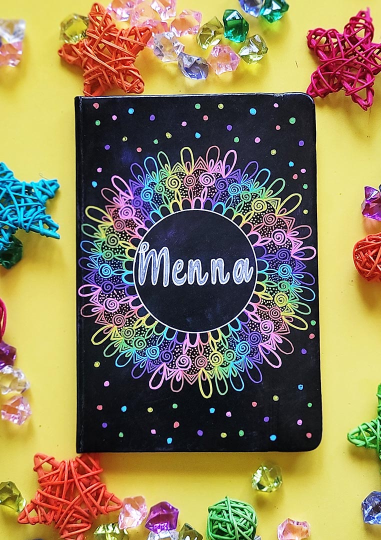 A Dark Sky hand painted Notebook with a mandala drawn in layers of pink then purple then blue then green then yellow then orange, with colored dots scattered all over and the name "Menna" written in silver in the center