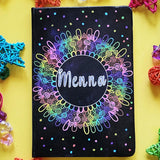 A Dark Sky hand painted Notebook with a mandala drawn in layers of pink then purple then blue then green then yellow then orange, with colored dots scattered all over and the name 