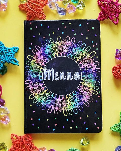 A Dark Sky hand painted Notebook with a mandala drawn in layers of pink then purple then blue then green then yellow then orange, with colored dots scattered all over and the name "Menna" written in silver in the center