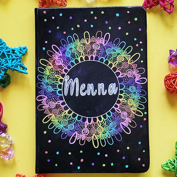 A Dark Sky hand painted Notebook with a mandala drawn in layers of pink then purple then blue then green then yellow then orange, with colored dots scattered all over and the name "Menna" written in silver in the center