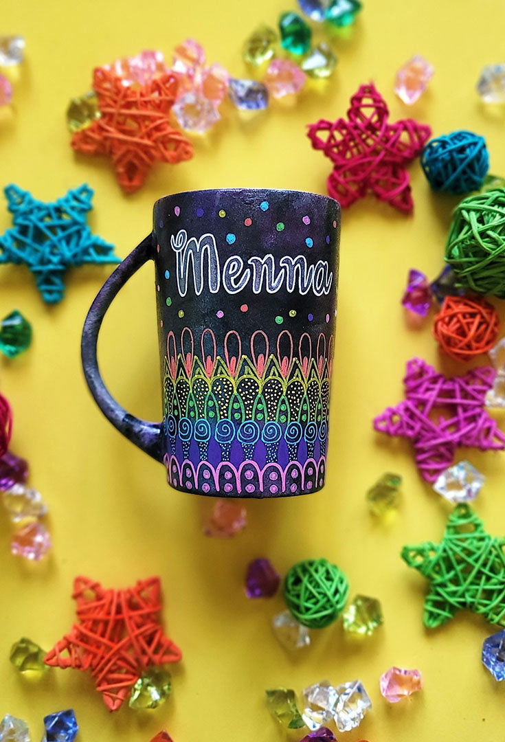 A Dark Sky hand painted mug with patterns on the bottom half horizontally inlayers of pink then purple then blue then green then yellow then orange, with colored dots scattered all over and the name "Menna" written in silver on the top