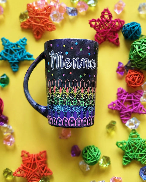 A Dark Sky hand painted mug with patterns on the bottom half horizontally inlayers of pink then purple then blue then green then yellow then orange, with colored dots scattered all over and the name "Menna" written in silver on the top