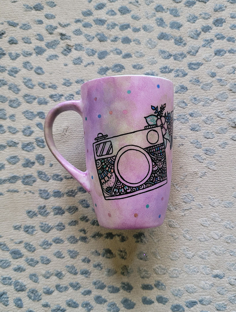 a pastel pink hand painted mug with a camera drawn on it with patterns inside and flowers coming out of its side