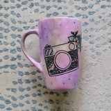 a pastel pink hand painted mug with a camera drawn on it with patterns inside and flowers coming out of its side