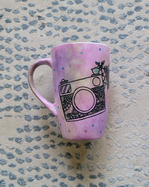 a pastel pink hand painted mug with a camera drawn on it with patterns inside and flowers coming out of its side