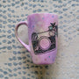 a pastel pink hand painted mug with a camera drawn on it with patterns inside and flowers coming out of its side