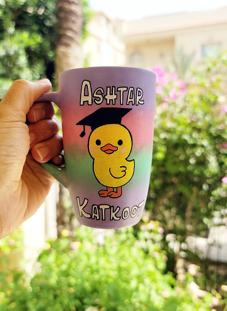 A gradient Hand painted mug (lilac=pink-mint-lilac) with the word "Ashtar" on top the the duck painted in the center wearing a graduation cap ,then the word "Katkoot" below it