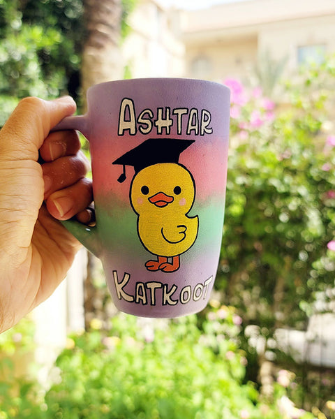 A gradient Hand painted mug (lilac=pink-mint-lilac) with the word "Ashtar" on top the the duck painted in the center wearing a graduation cap ,then the word "Katkoot" below it