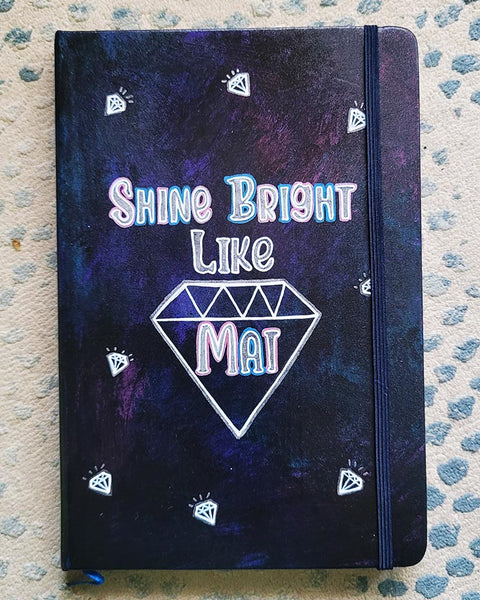 A Dark Sky hand painted notebook with the words shine bright like and a diamond drawn with the name "Mai" inside