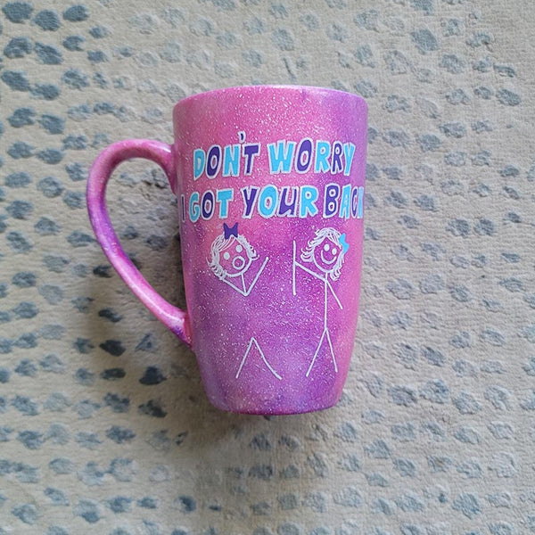 A hand painted pink galaxy mug with the words "don't worry, I got your back " and a drawing of 2 stick figures with the girl holding the back of the other girl