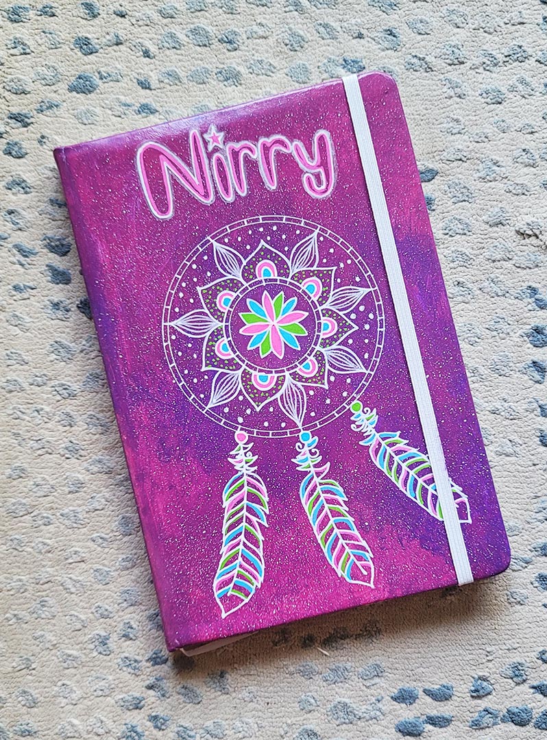 A Pink galaxy hand painted notebook with a dreamcatcher drawn in white and the name "Nirry" written in the top center