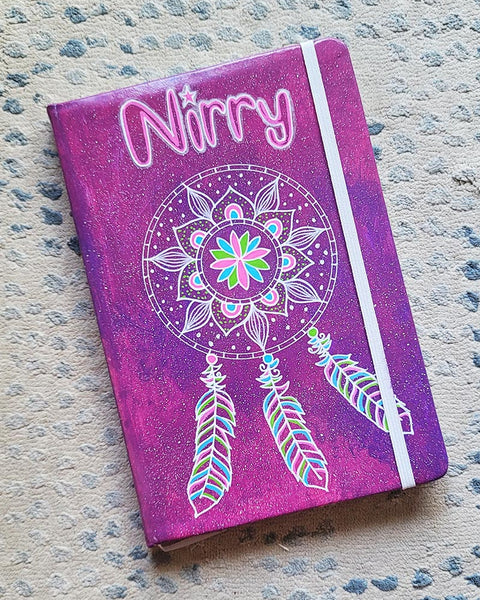 A Pink galaxy hand painted notebook with a dreamcatcher drawn in white and the name "Nirry" written in the top center