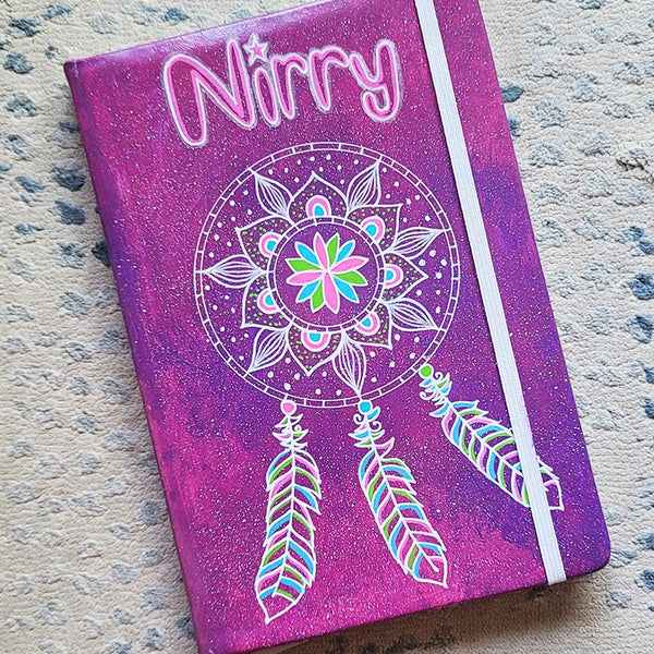 A Pink galaxy hand painted notebook with a dreamcatcher drawn in white and the name "Nirry" written in the top center