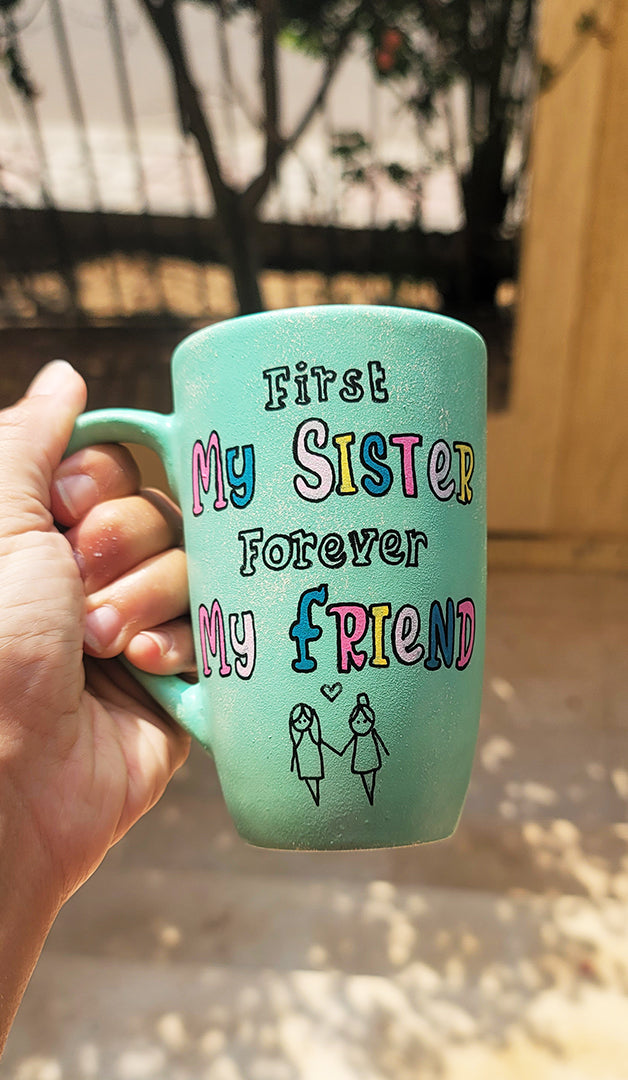 a mint green hand painted mug with the words "first my sister, forever my friend" and a sketch of 2 girl stick figures holding hands