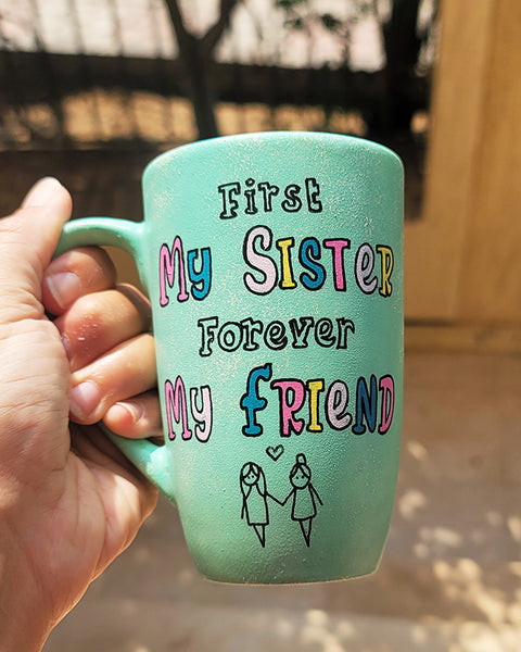 a mint green hand painted mug with the words "first my sister, forever my friend" and a sketch of 2 girl stick figures holding hands