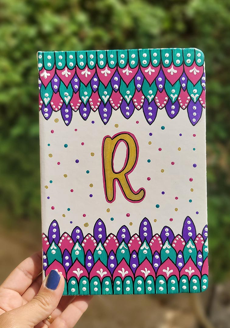 A Pearl white notebook with patterns on both top and bottom in colors of pink, turquoise and purple and the letter R in the center in gold with dots of pink, purple, gold and turquoise scattered all over