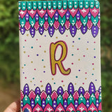 A Pearl white notebook with patterns on both top and bottom in colors of pink, turquoise and purple and the letter R in the center in gold with dots of pink, purple, gold and turquoise scattered all over