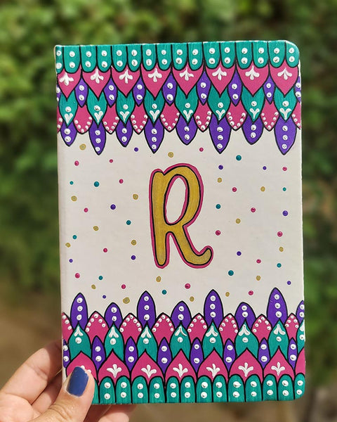 A Pearl white notebook with patterns on both top and bottom in colors of pink, turquoise and purple and the letter R in the center in gold with dots of pink, purple, gold and turquoise scattered all over