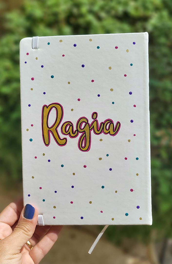 A Pearl white notebook with the name "ragia" in the center adn dots of purple, pink, gold and turquoise scattered all over