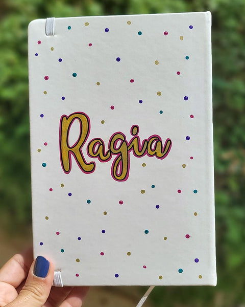 A Pearl white notebook with the name "ragia" in the center adn dots of purple, pink, gold and turquoise scattered all over