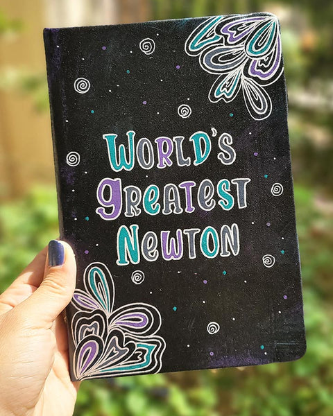 A Black galaxy hand painted notebook with doodles coming from 2 side corners and the words "World's greartest newton" in the center with small swirls scattered all over