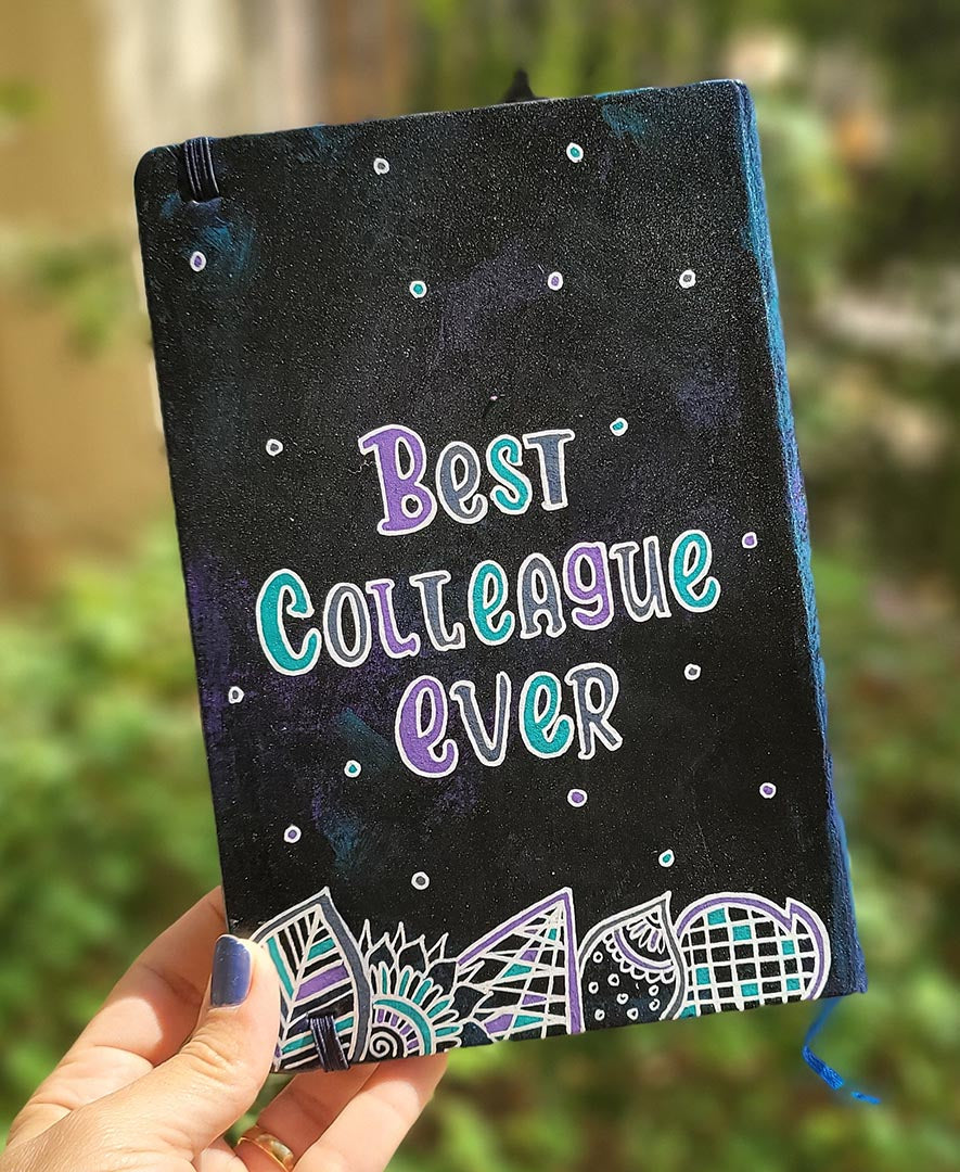 A black galaxy hand painted notebook with doodles at the bottom and the words "Best Colleague Ever" written in the center