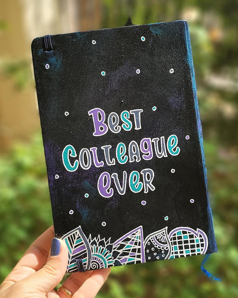 A black galaxy hand painted notebook with doodles at the bottom and the words "Best Colleague Ever" written in the center