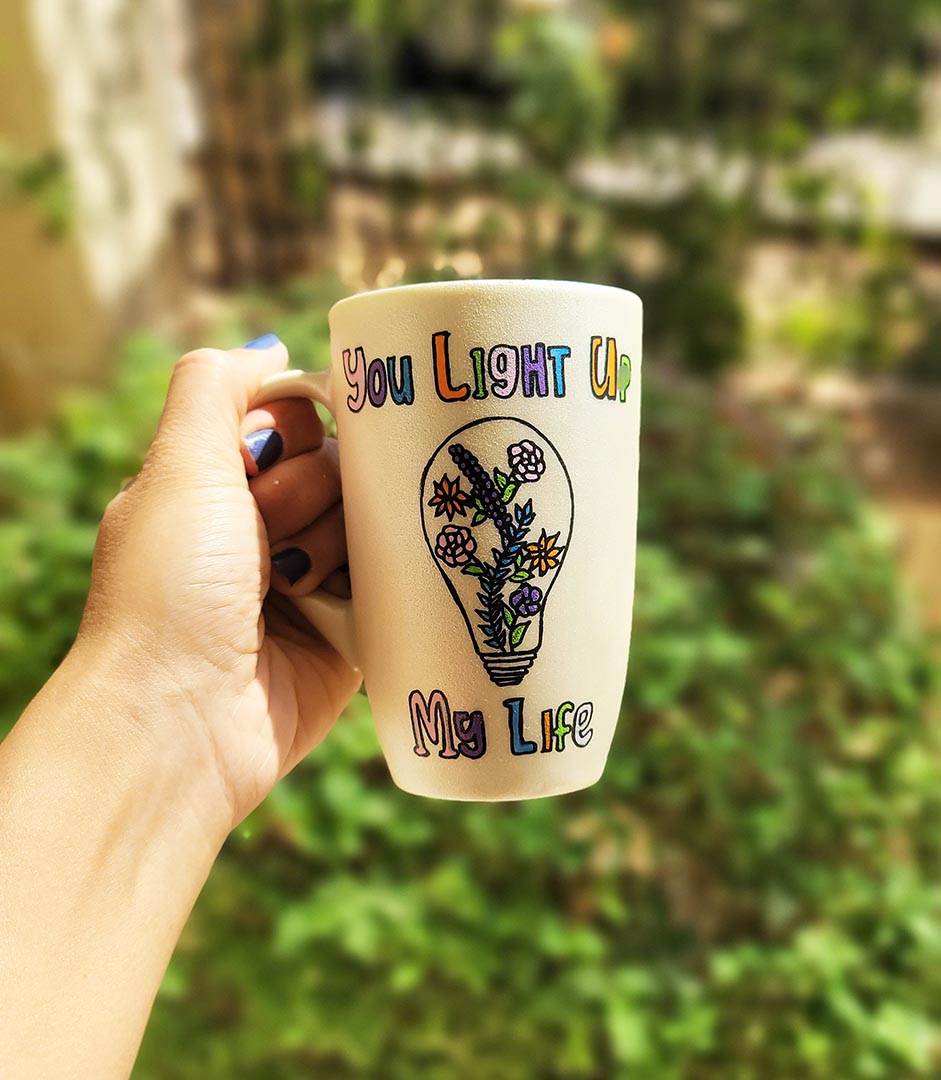 A pearl white hand-painted mug with the words "you light up my life" and a bulb drawn on it with flowers inside