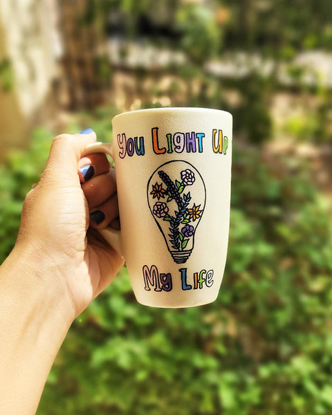 A pearl white hand-painted mug with the words "you light up my life" and a bulb drawn on it with flowers inside