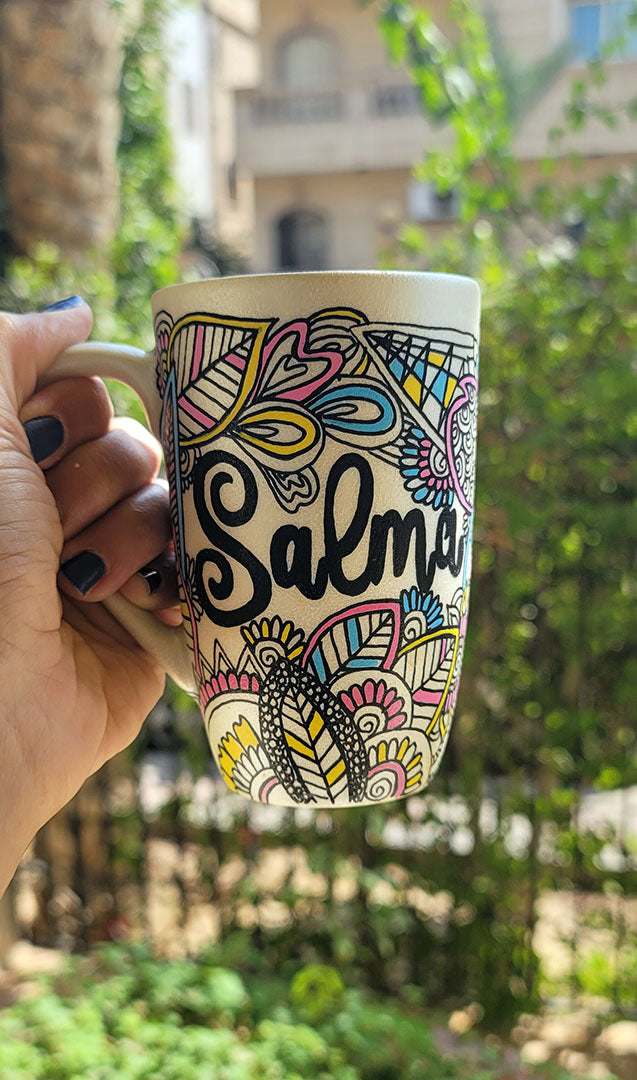 A Pearl white hand painted mug with dooldes drawn all over in black and colored in pinks, yellow and blue, with the name "Salma" written in between