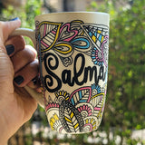 A Pearl white hand painted mug with dooldes drawn all over in black and colored in pinks, yellow and blue, with the name 