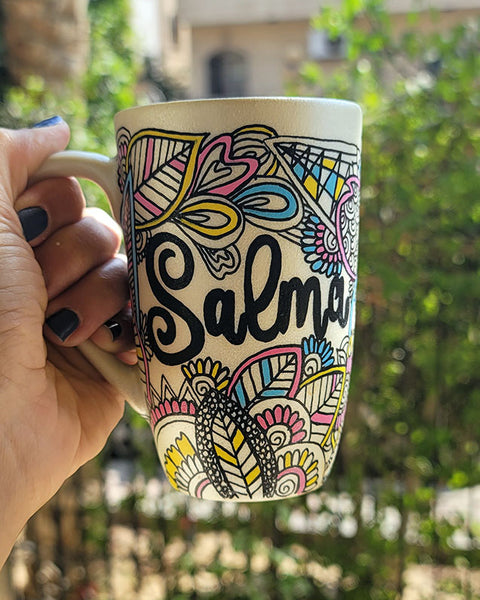 A Pearl white hand painted mug with dooldes drawn all over in black and colored in pinks, yellow and blue, with the name "Salma" written in between