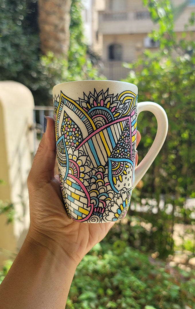 A Pearl white hand painted mug with dooldes drawn all over in black and colored in pinks, yellow and blue