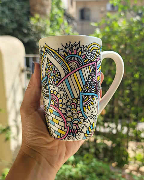 A Pearl white hand painted mug with dooldes drawn all over in black and colored in pinks, yellow and blue