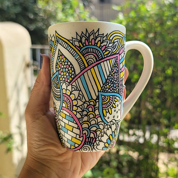 A Pearl white hand painted mug with dooldes drawn all over in black and colored in pinks, yellow and blue