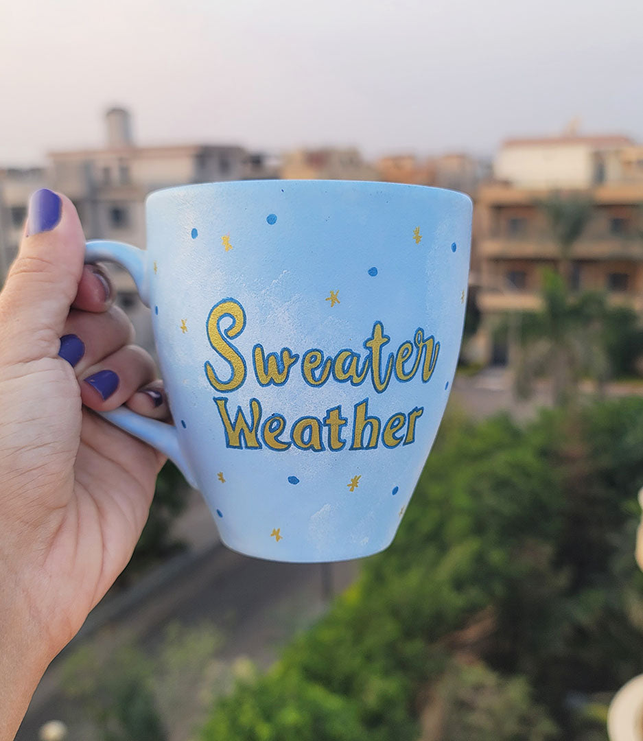 A baby blue hand painted mug with the sentence "Sweater Weather" in gold