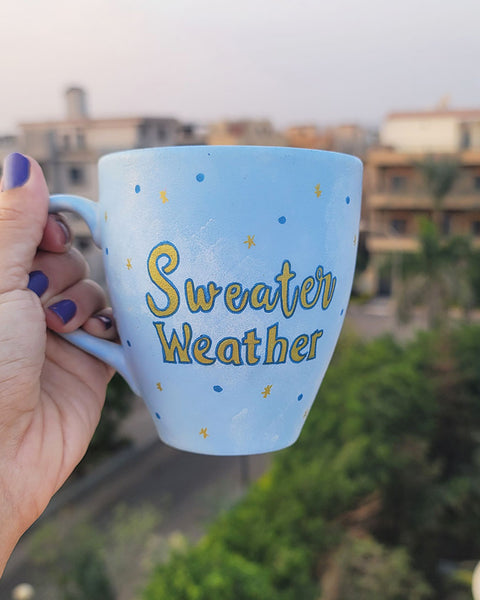 A baby blue hand painted mug with the sentence "Sweater Weather" in gold