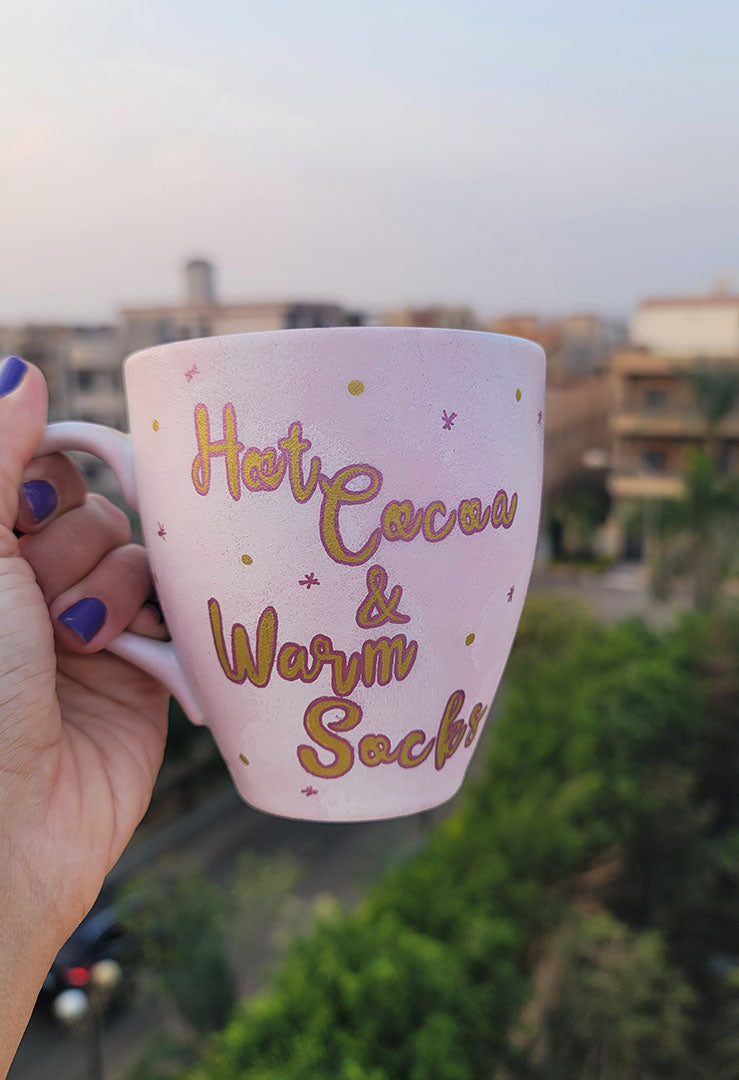 A Hand painted Baby Pink mug with the sentence "Hot Cocoa and warm socks" in gold