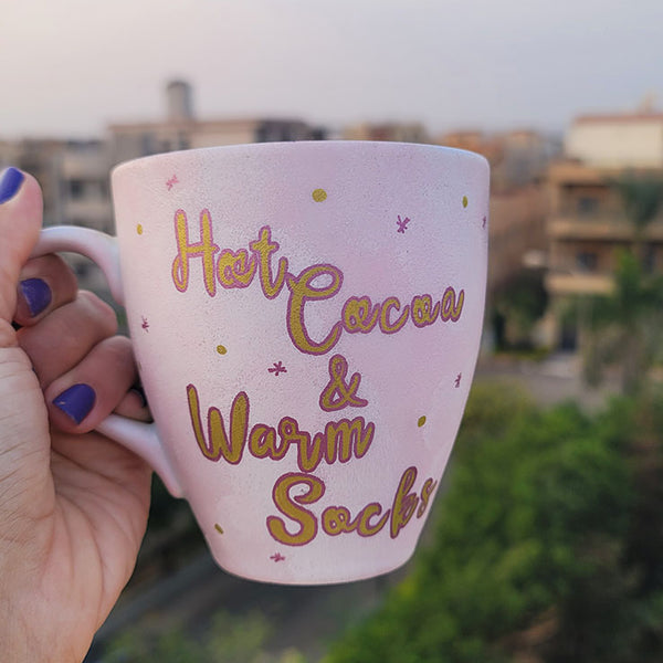 A Hand painted Baby Pink mug with the sentence "Hot Cocoa and warm socks" in gold