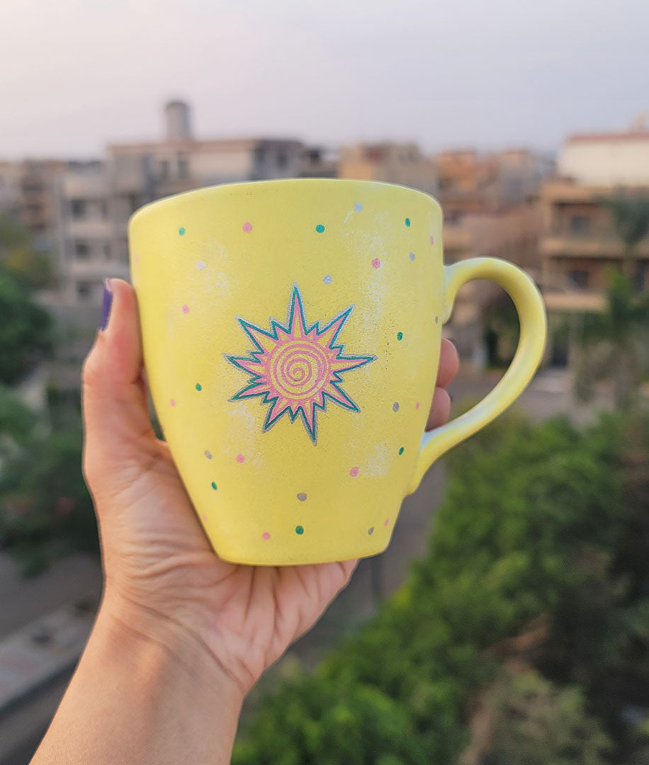 A Pastel yellow hand painted mug with A small drawing of a sun in pink and turquoise