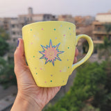 A Pastel yellow hand painted mug with A small drawing of a sun in pink and turquoise