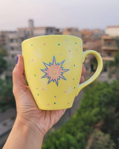 A Pastel yellow hand painted mug with A small drawing of a sun in pink and turquoise