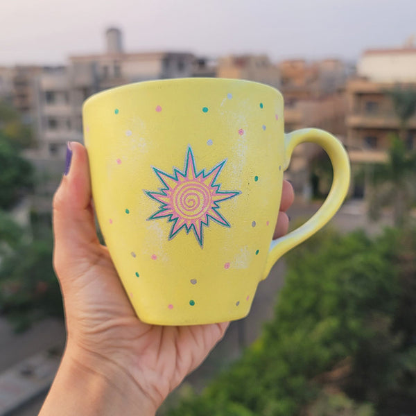 A Pastel yellow hand painted mug with A small drawing of a sun in pink and turquoise